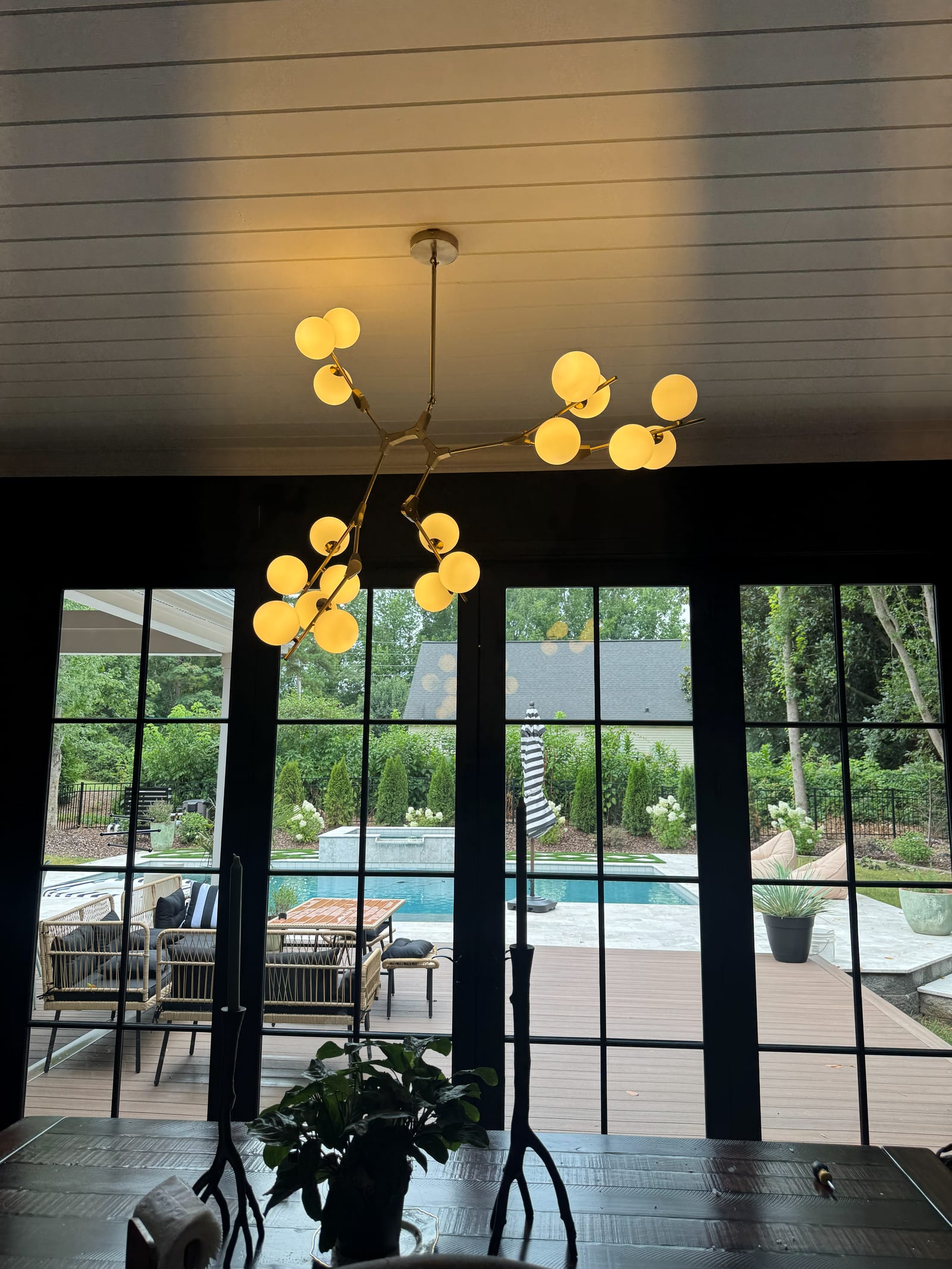 Brass branch chandelier with frosted globe bulbs hanging above a dining table, in front of black steel french doors looking out to a pool