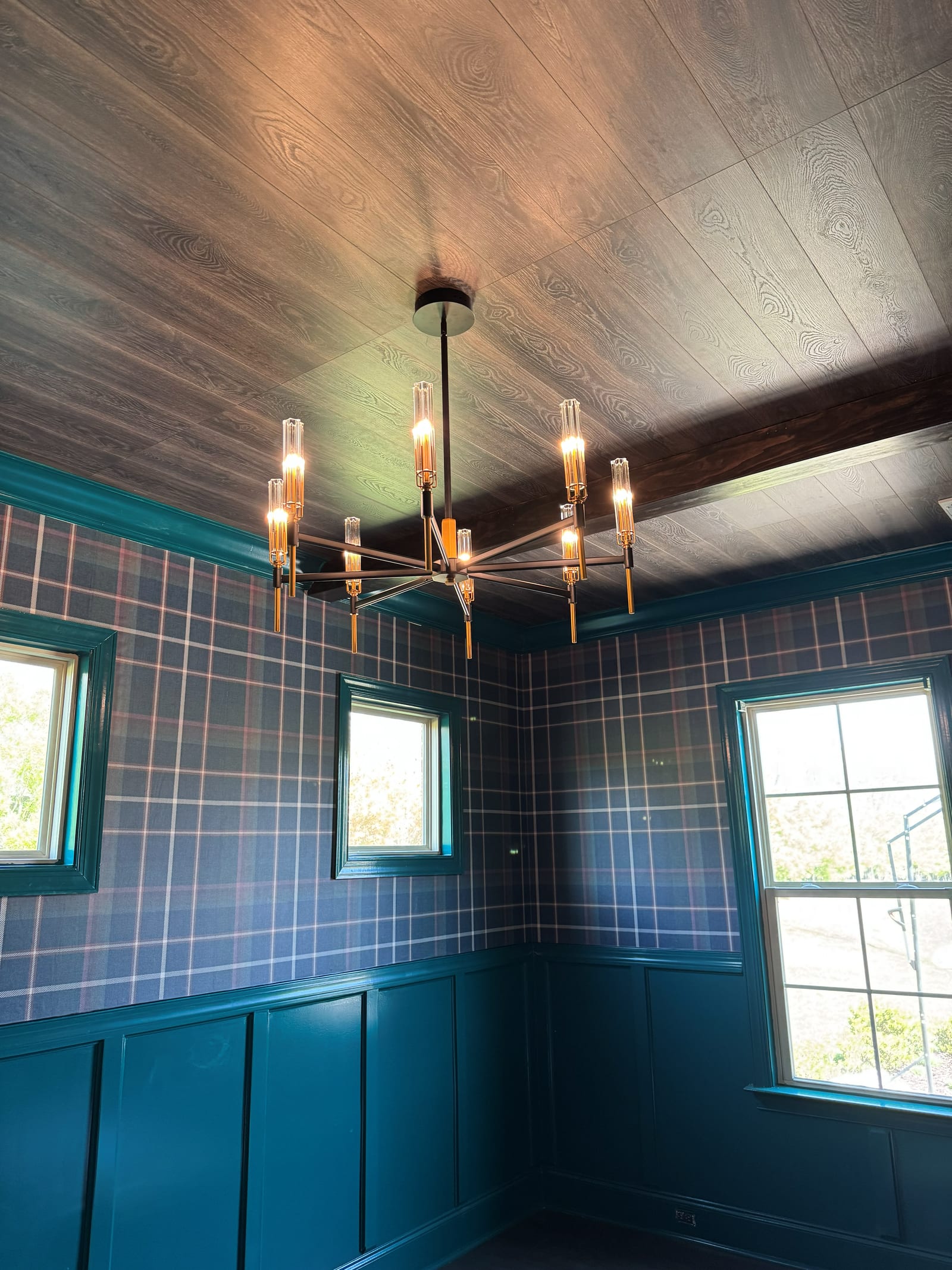 Modern brass and black chandelier hanging from a wood plank ceiling in a room with teal panelling and plaid wallpaper