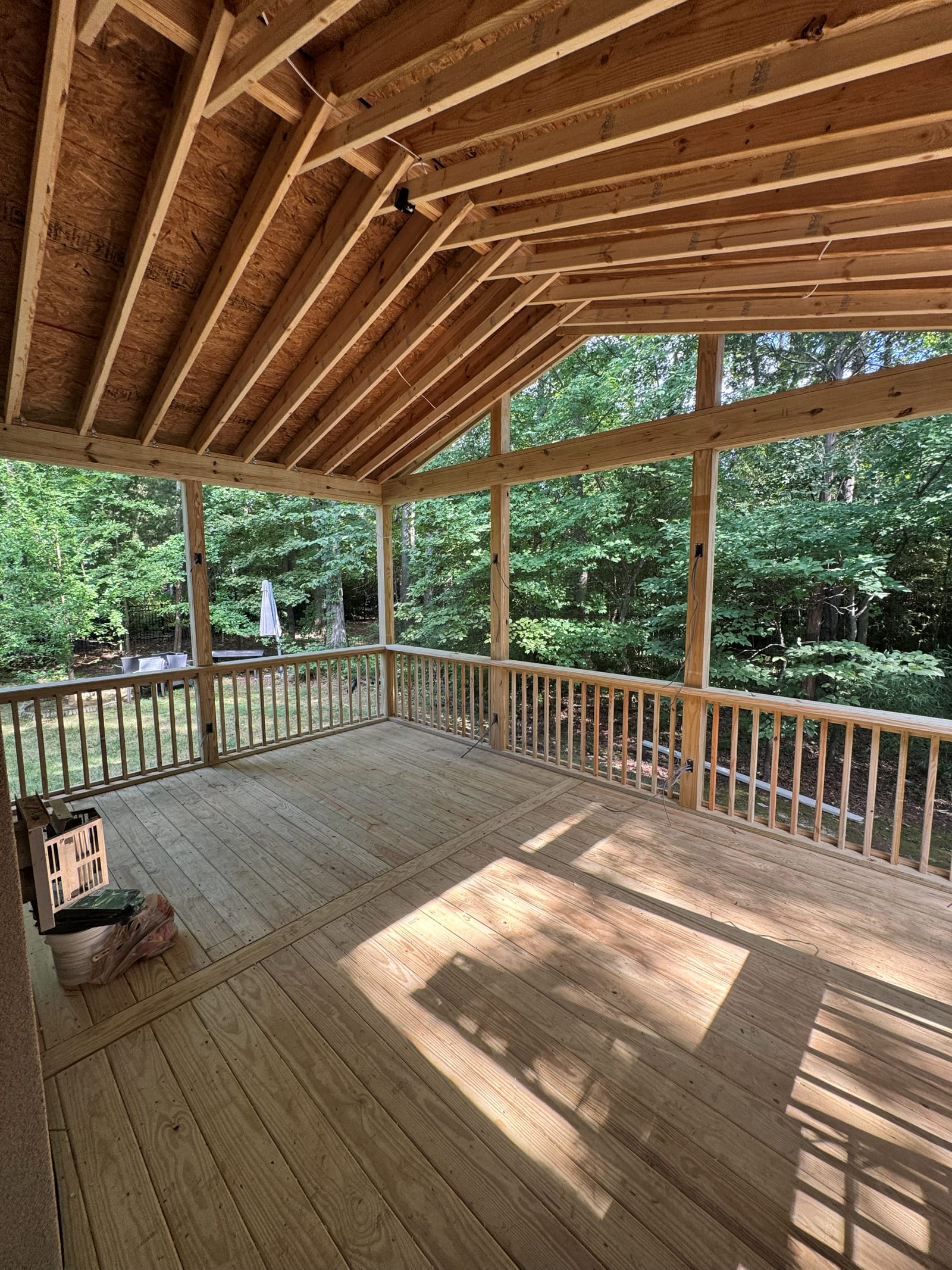 Vaulted screened porch under construction with exposed rafters, roughed-in wiring runs and a fan box in the ceiling, looking out to woods